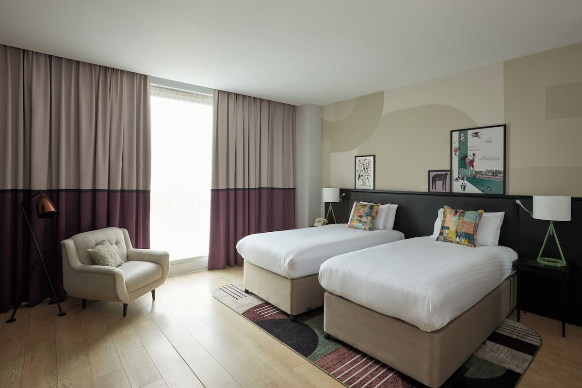 Quarto de hotel com duas camas, uma cadeira, uma luminária e uma obra de arte na parede.