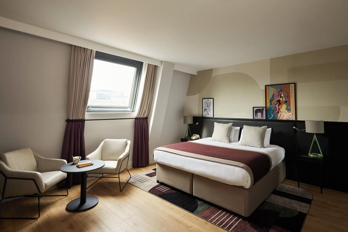 Quarto de hotel com cama, duas cadeiras, uma mesinha, mesa de cabeceira e janela com cortinas.