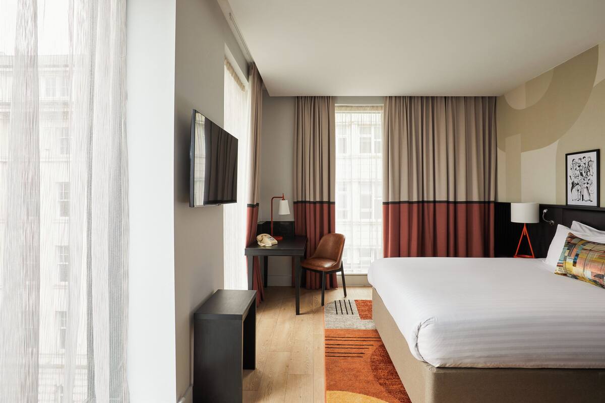 Quarto de hotel com cama, escrivaninha, cadeira e televisão fixada na parede.