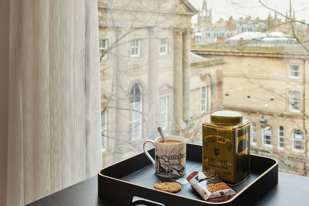 A tray with a mug, a tea tin, and a biscuit on a table by a window overlooking a historic building.