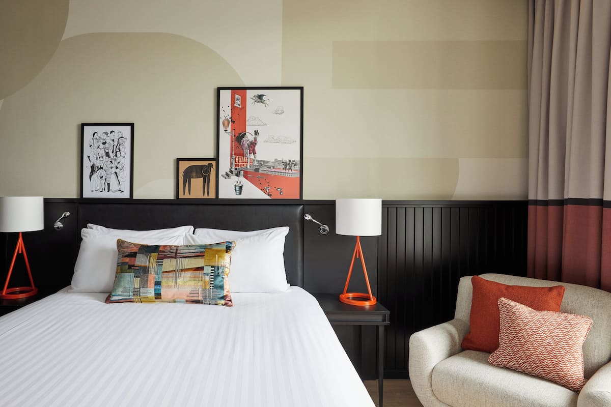 Quarto de hotel com uma cama grande, duas mesas de cabeceira, uma cadeira e obras de arte na parede.