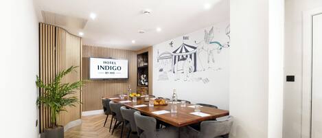 A modern hotel lobby with a wooden reception desk, a large wall mural, and a dining area with a long wooden table and grey chairs.