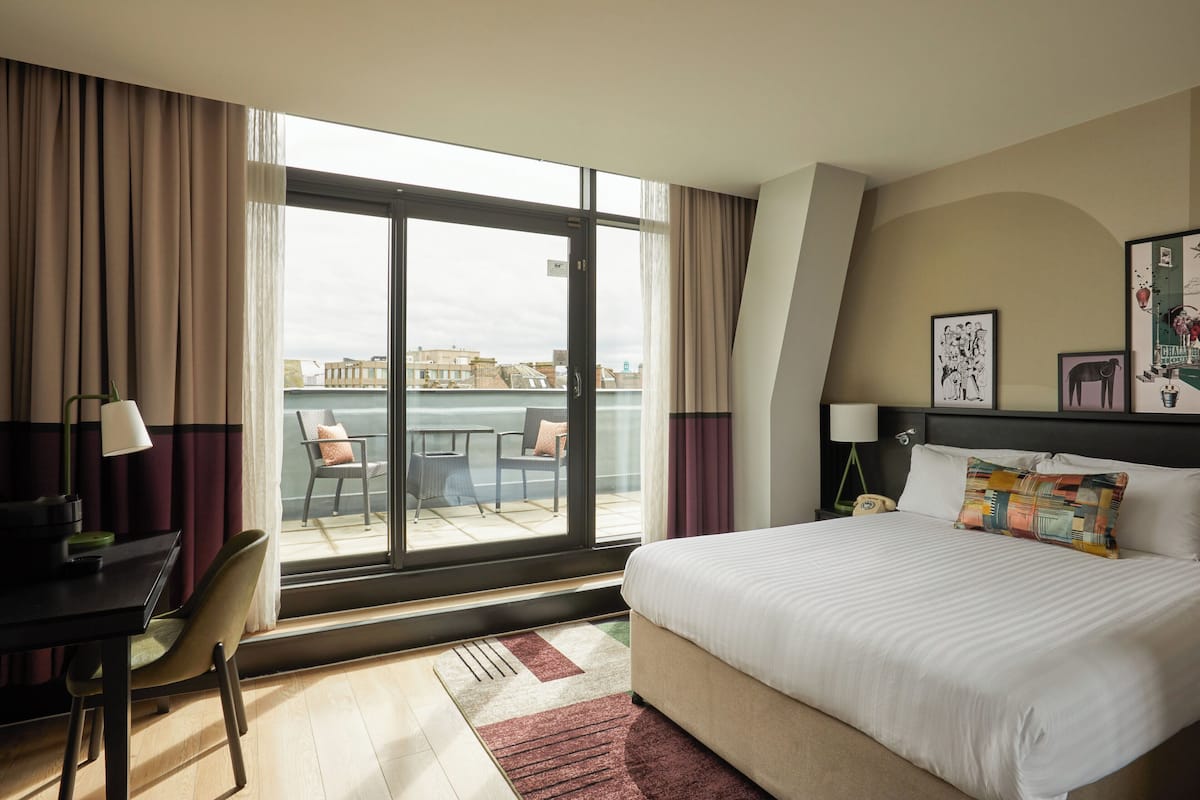 Quarto de hotel com uma cama grande, uma escrivaninha com abajur, uma cadeira e vista para a cidade através de uma janela ampla.