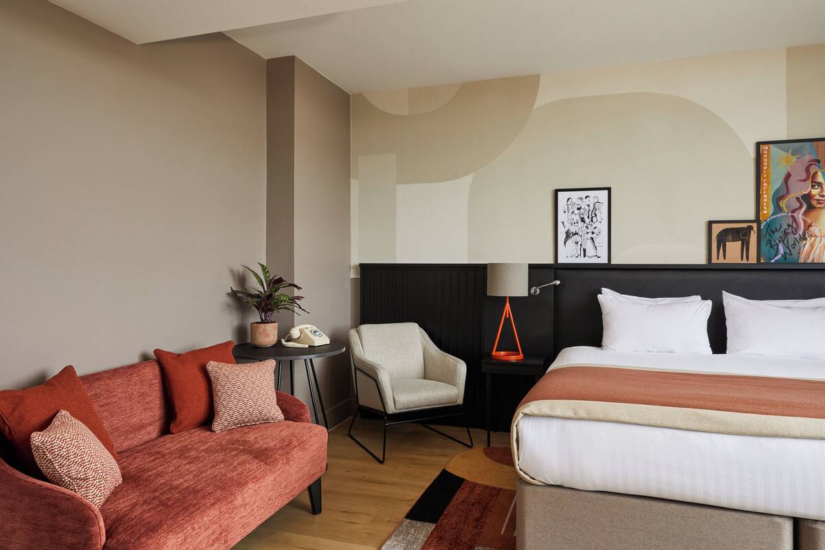 Quarto de hotel com uma cama, um sofá, uma cadeira, uma mesinha com abajur e obras de arte na parede.