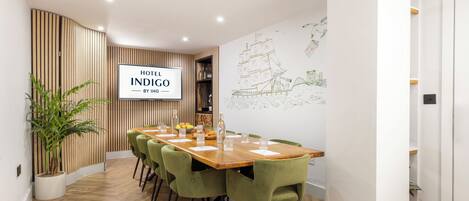 A modern hotel room with a wooden floor, a dining table with green chairs, and a wall-mounted sign reading "HOTEL INDIGO BY IHG".