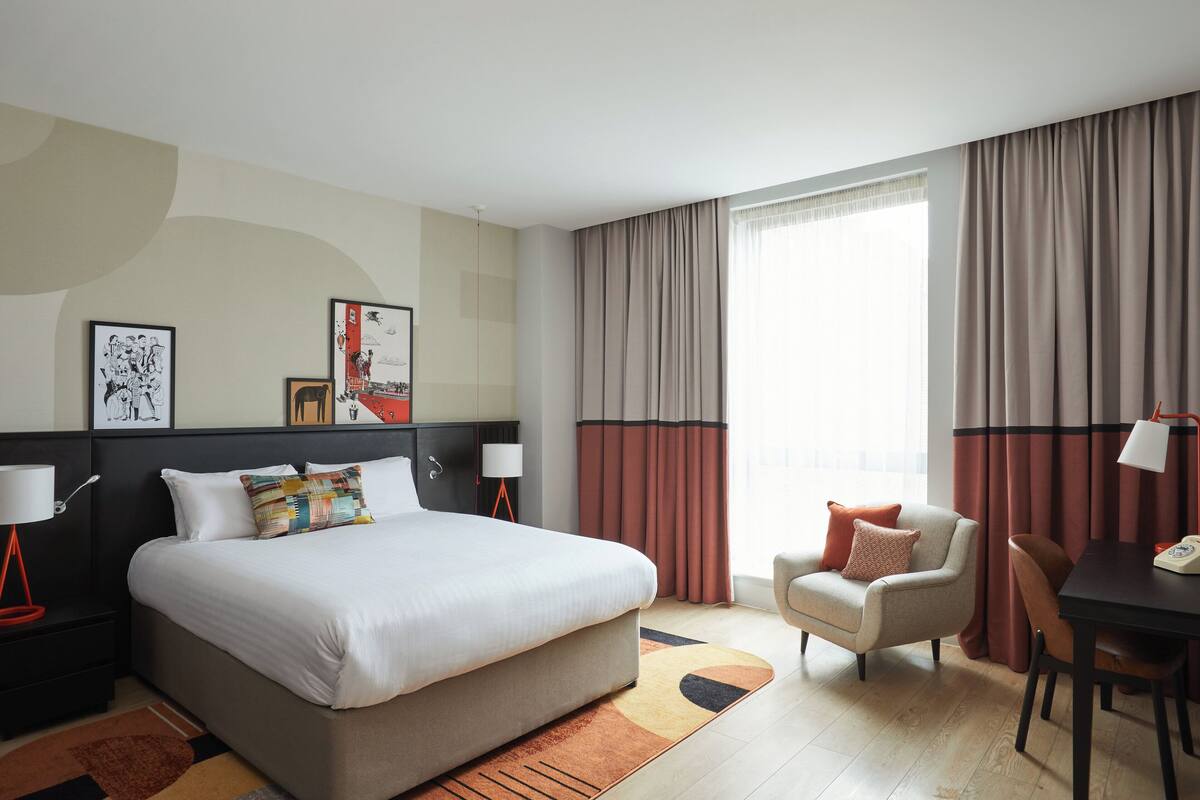 Quarto de hotel moderno com uma cama grande, uma escrivaninha, uma cadeira e um sofá. Há cortinas, uma luminária e obras de arte nas paredes.