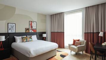 A modern hotel room with a large bed, a desk, a chair, and a sofa. There are curtains, a lamp, and artwork on the walls.