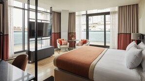 A hotel room with a large bed, a desk, a chair, a small table, and a TV. The room has large windows with curtains and a view of the water.