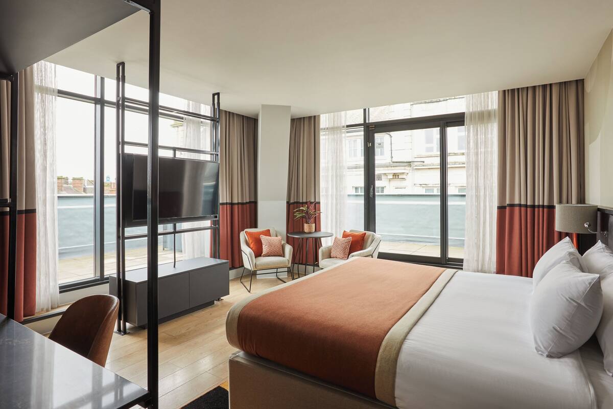 Quarto de hotel com uma cama grande, escrivaninha, cadeira, mesa pequena e televisão. O quarto possui janelas amplas com cortinas e vista para o mar.