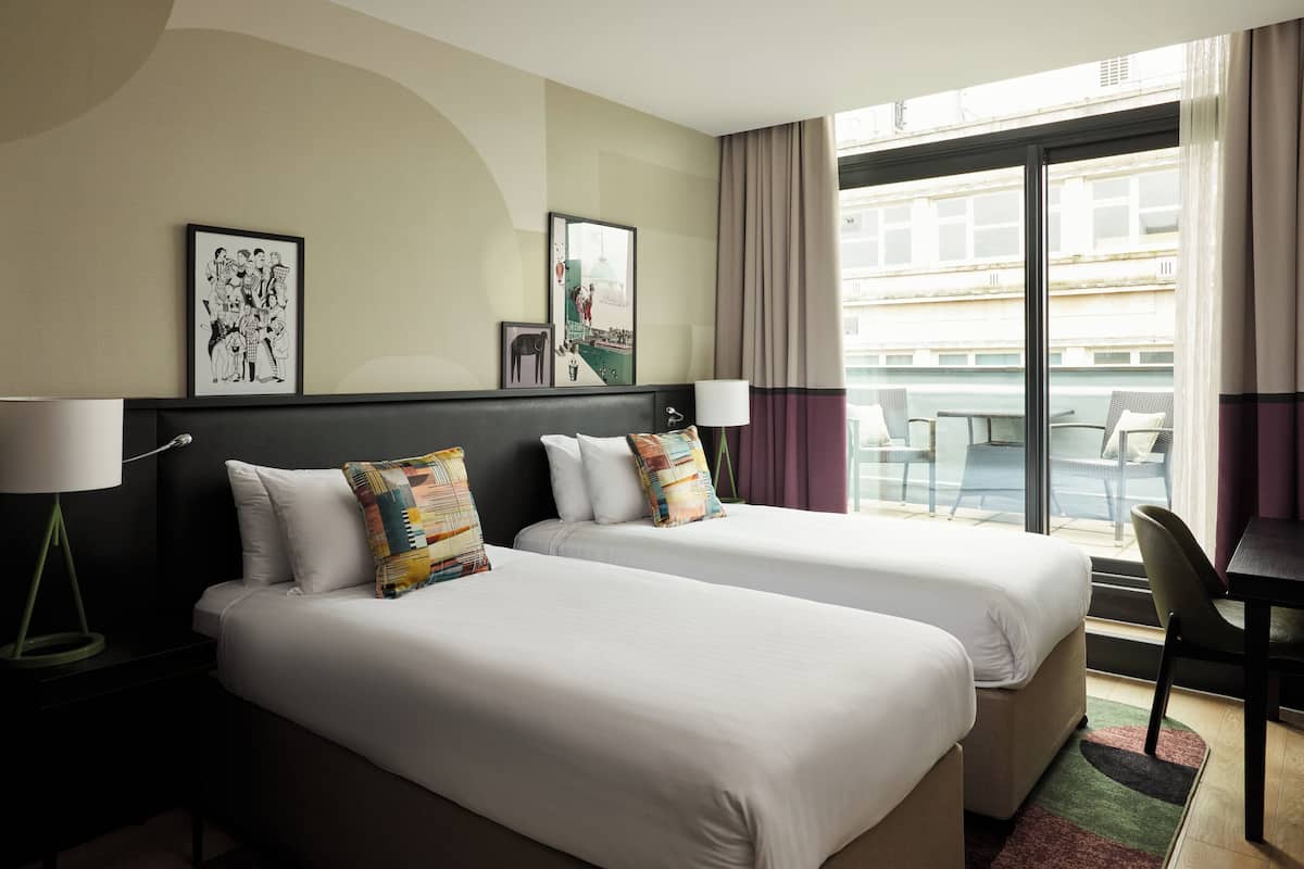 Quarto de hotel com duas camas, uma mesa e uma cadeira. Há duas fotos emolduradas na parede e uma janela com cortinas.
