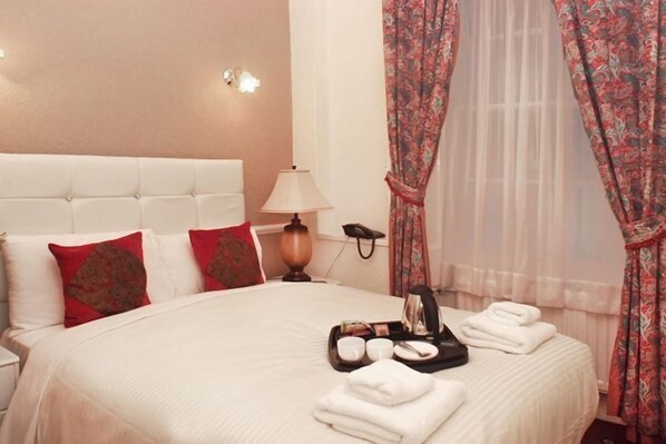A hotel room with a large bed, white headboard, red and brown pillows, a bedside lamp, a telephone, and a tray with towels and slippers. - Classic Hyde Park Hotel (London)