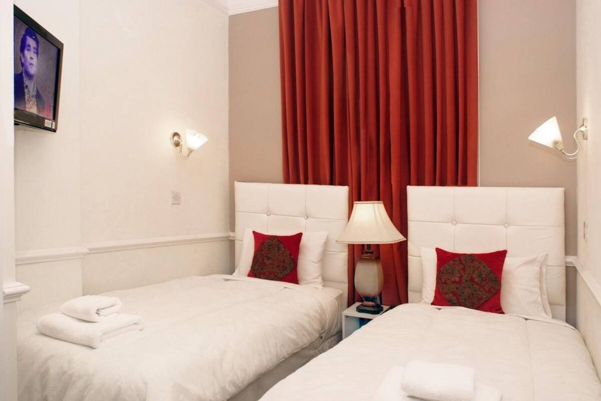 Une chambre d’hôtel avec deux lits, chacun étant équipé de draps blancs et d’oreillers rouges. Un téléviseur est fixé au mur, une lampe se trouve sur la table de chevet, et il y a un rideau rouge.
