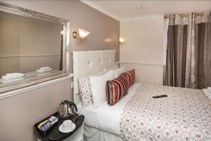 A hotel room with a large mirror, a bed with white bedding and floral-patterned covers, a bedside table with a tray of toiletries, and a window with curtains. - Classic Hyde Park Hotel (London)