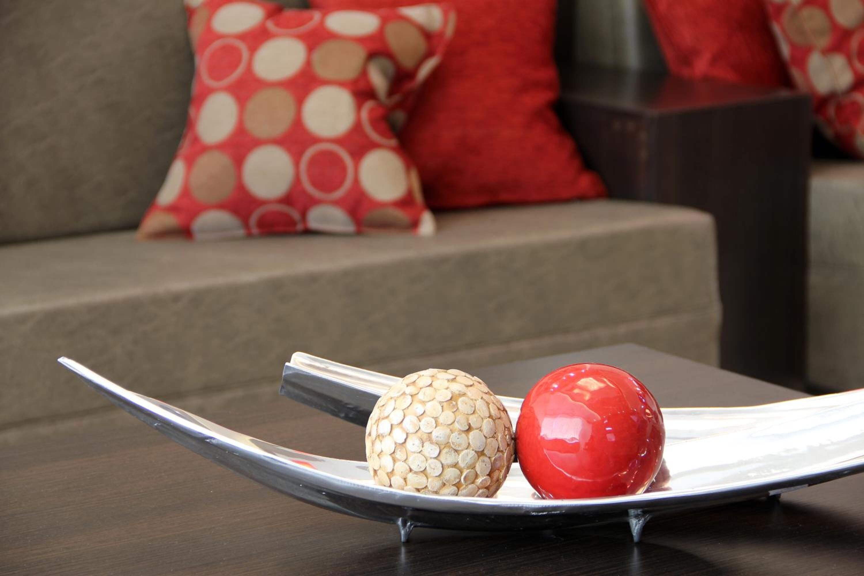 Una mesa con una bandeja metálica que sostiene dos objetos esféricos, una esfera con textura y una esfera roja brillante, sobre una mesa de madera con un sofá al fondo.