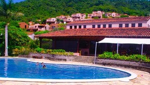 Una piscina con plataforma de baldosas, rodeada de vegetación exuberante y una colina con edificios tradicionales.