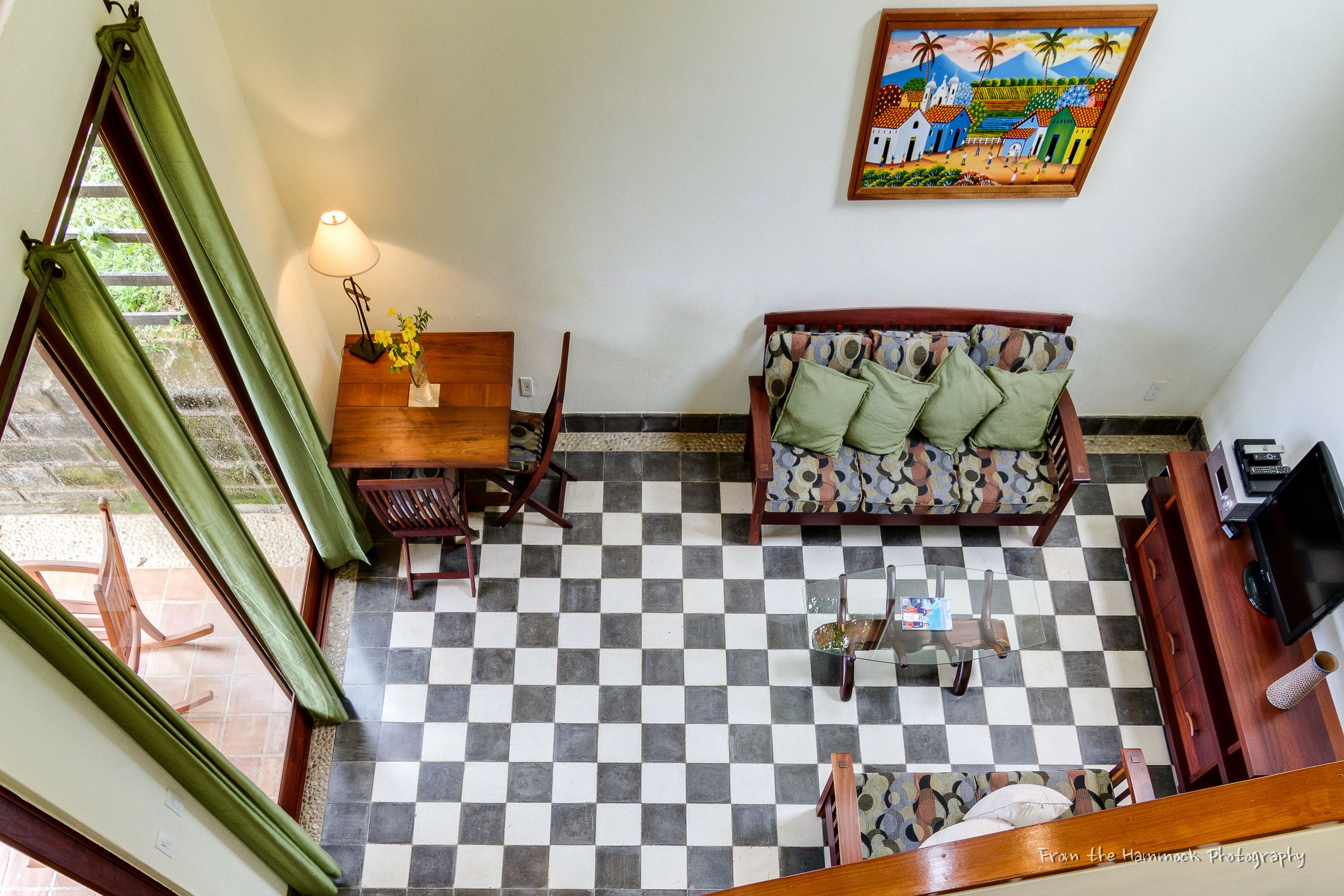 Piso xadrez, um sofá com almofadas estampadas, uma mesa de cabeceira de madeira com uma luminária e um quadro na parede.