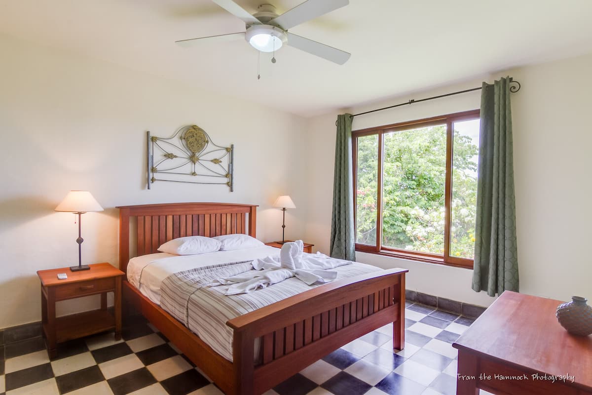 Ein Schlafzimmer mit kariertem Boden, einem hölzernen Bett, zwei Nachttischen mit Lampen, einem Deckenventilator, einem Fenster mit Vorhängen und Blick ins Grüne.