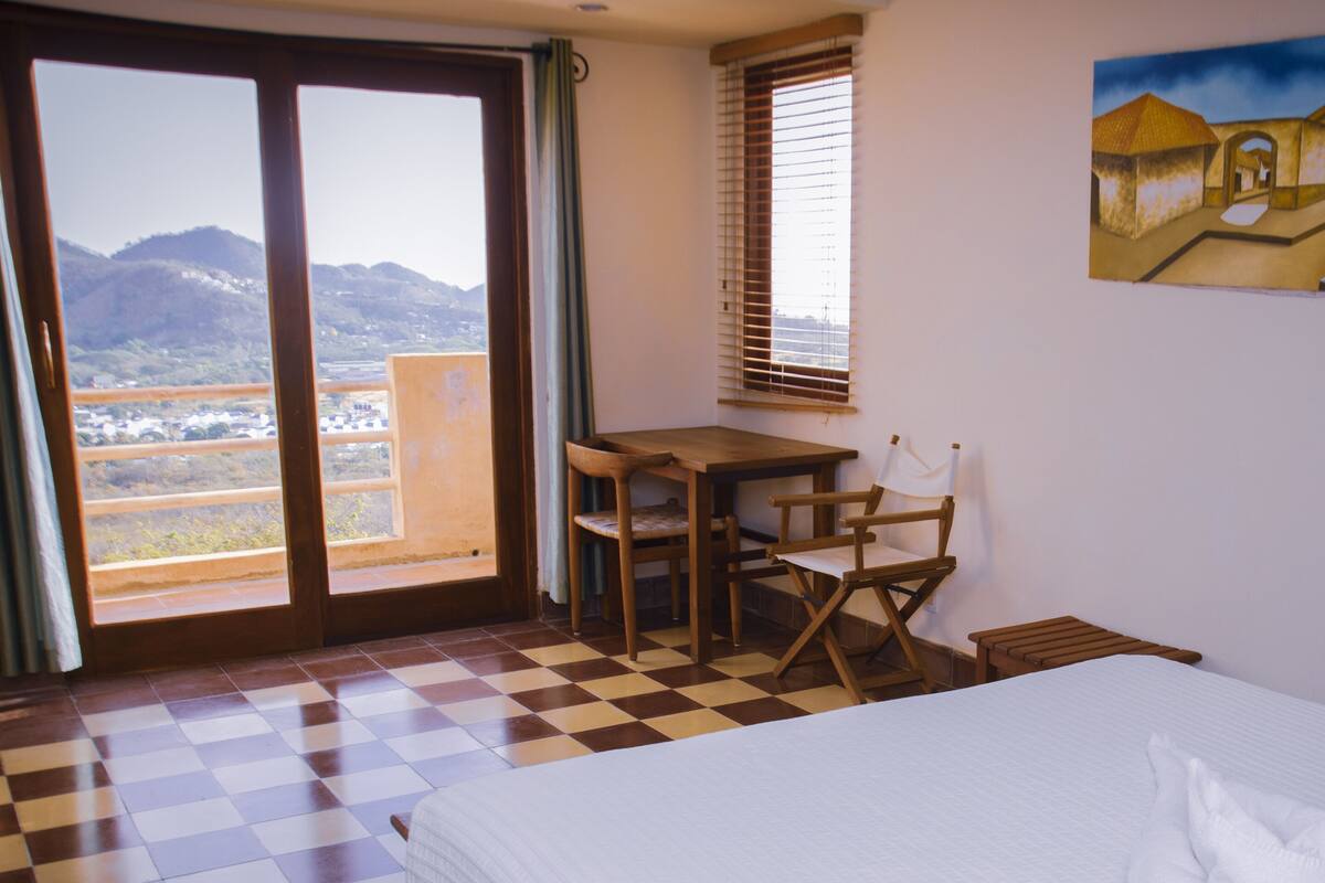 Ein Hotelzimmer mit einem Bett, einem Schreibtisch und einem Stuhl. An der Wand hängt ein Gemälde und der Blick geht auf die Berge durch das Fenster.