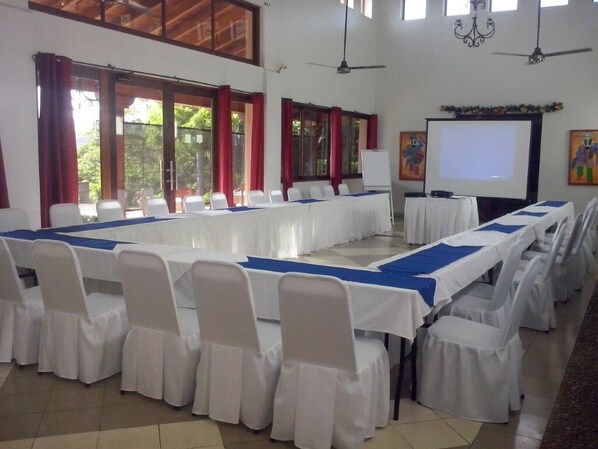 A conference room with long tables and white chairs, large windows, and a projector screen.