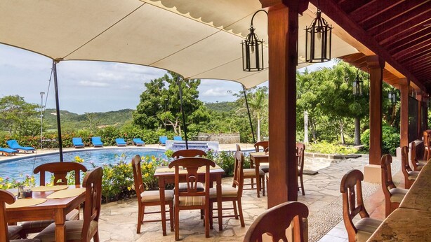 Un espacio de estar al aire libre techado con mesas y sillas de madera, con vistas a una piscina y abundante vegetación.