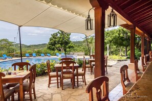 A covered outdoor seating area with wooden tables and chairs, overlooking a swimming pool and lush greenery.