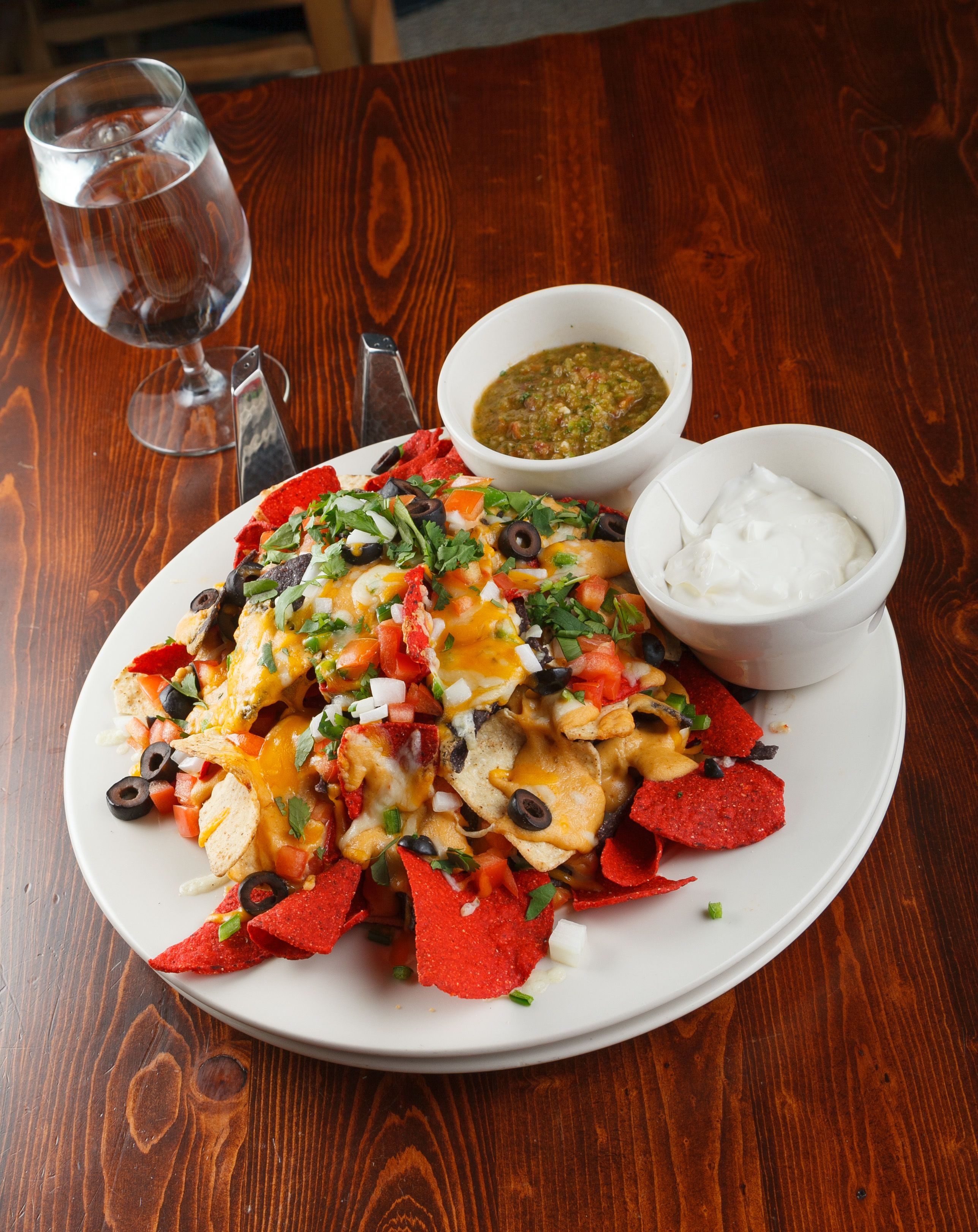 Prato de nachos com queijo, azeitonas pretas e cobertura verde, acompanhado de um recipiente com creme azedo e um molho de salsa verde.