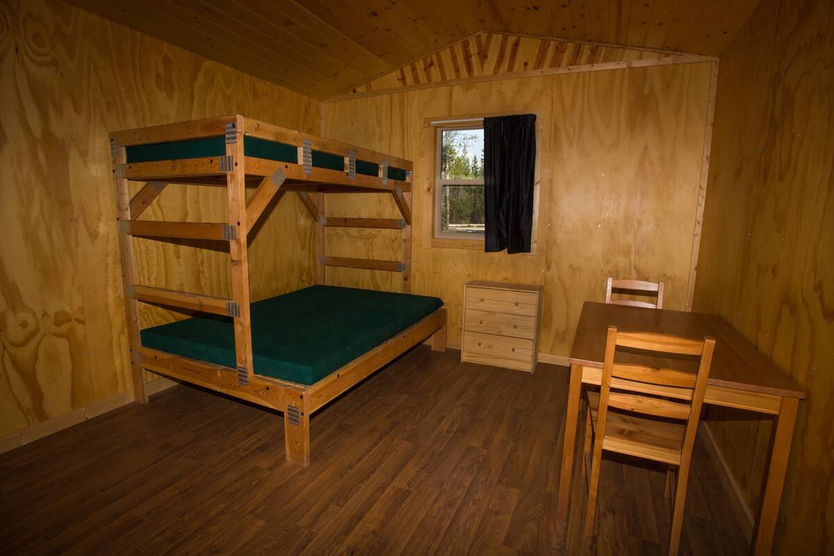 Un lit superposé en bois avec un matelas vert, un bureau en bois avec des tiroirs et une fenêtre avec un rideau noir.