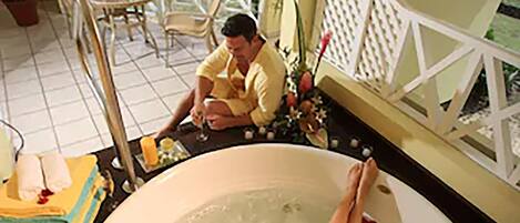 A person relaxing in a large bathtub filled with water and rose petals, with a view of the ocean and palm trees in the background.