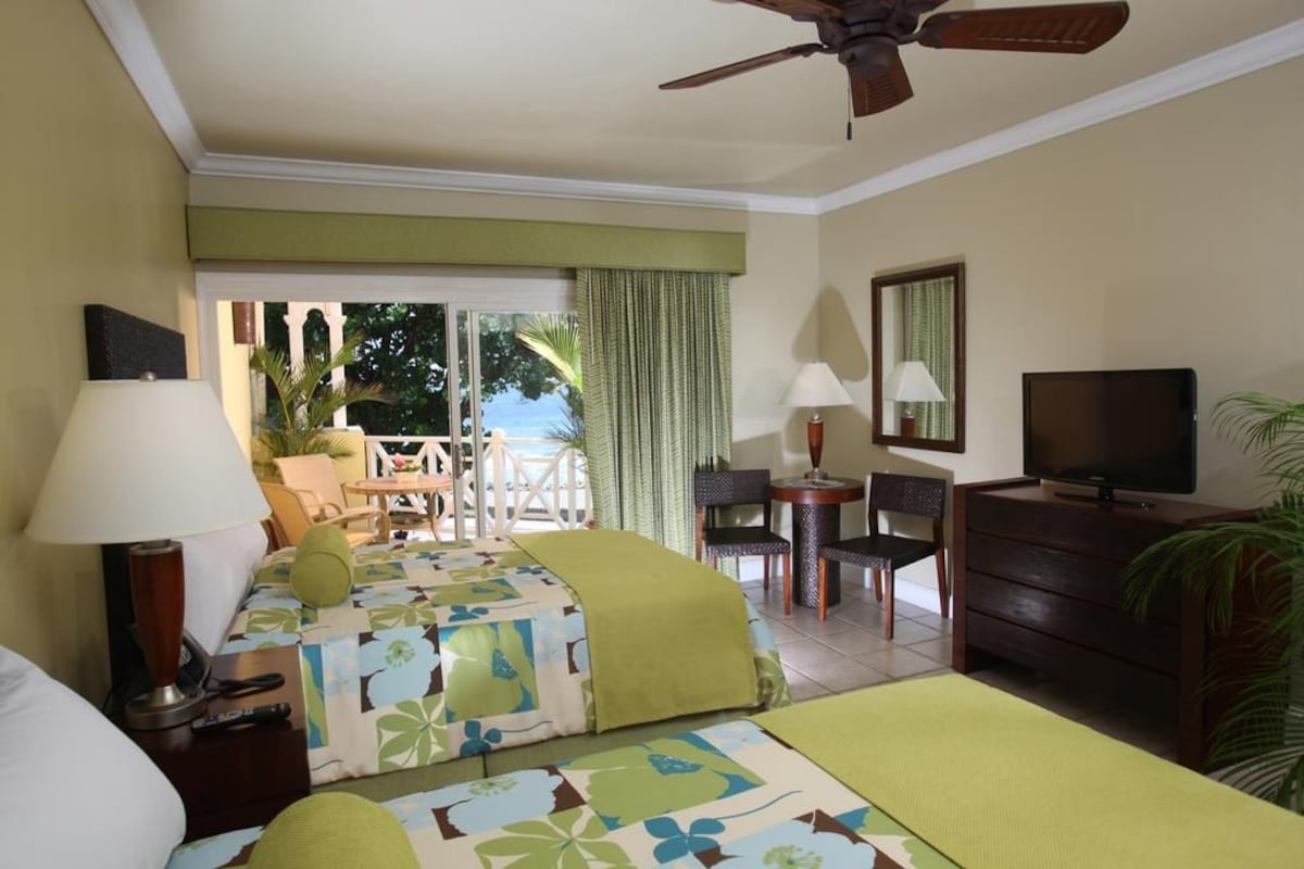 A hotel room with two beds, a desk, a chair, a TV, and a ceiling fan.