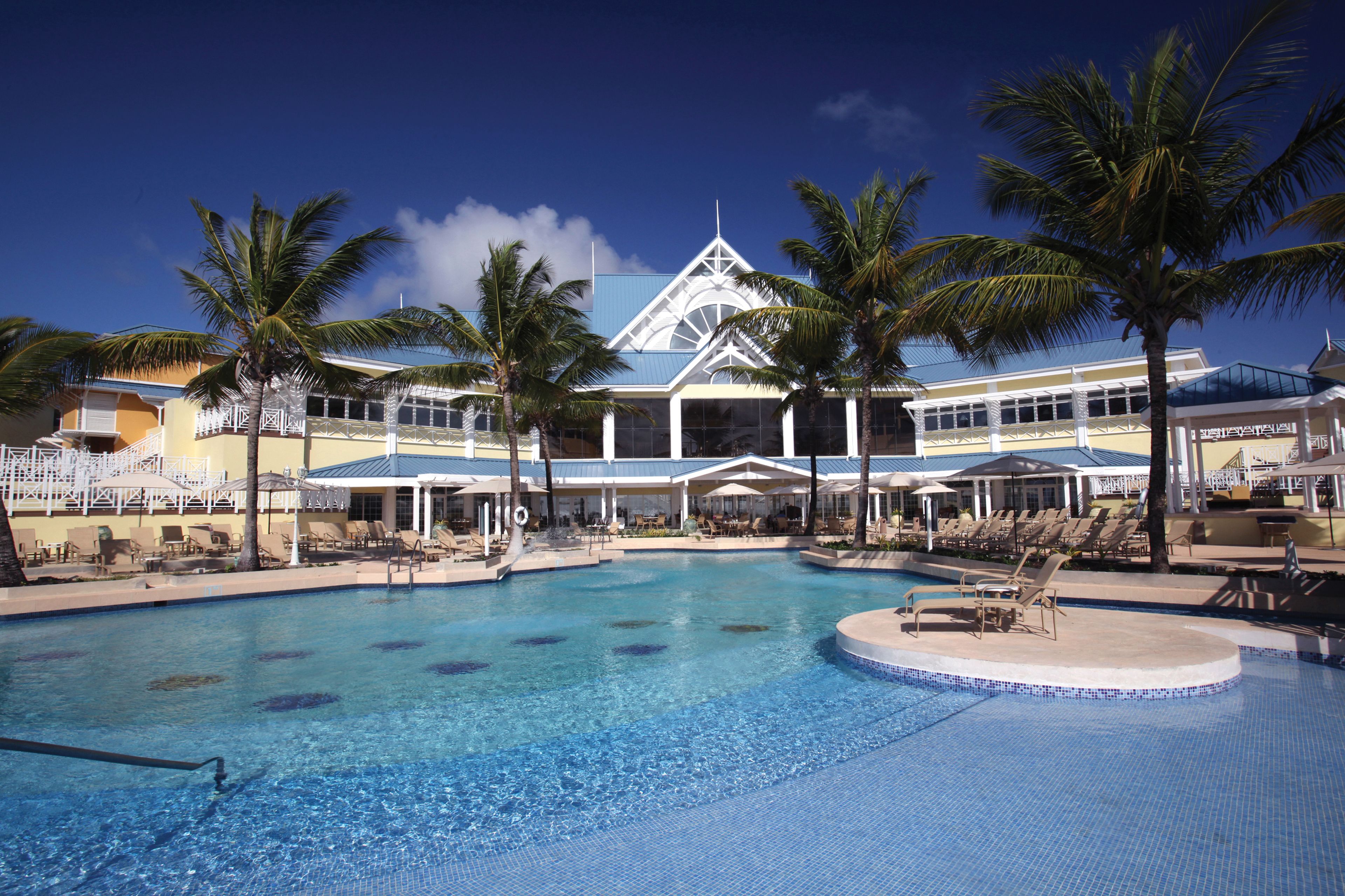 A swimming pool with lounge chairs and palm trees, surrounded by a building with a blue roof and white columns.