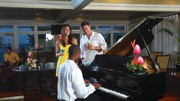 A grand piano in a well-lit room with a large window, a dining area with a round table, and a bar area with a bartender.