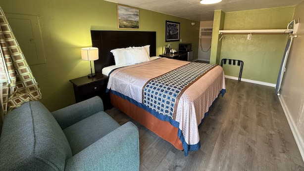 A hotel room with a bed, a desk, a chair, a lamp, a painting, and a shower area.