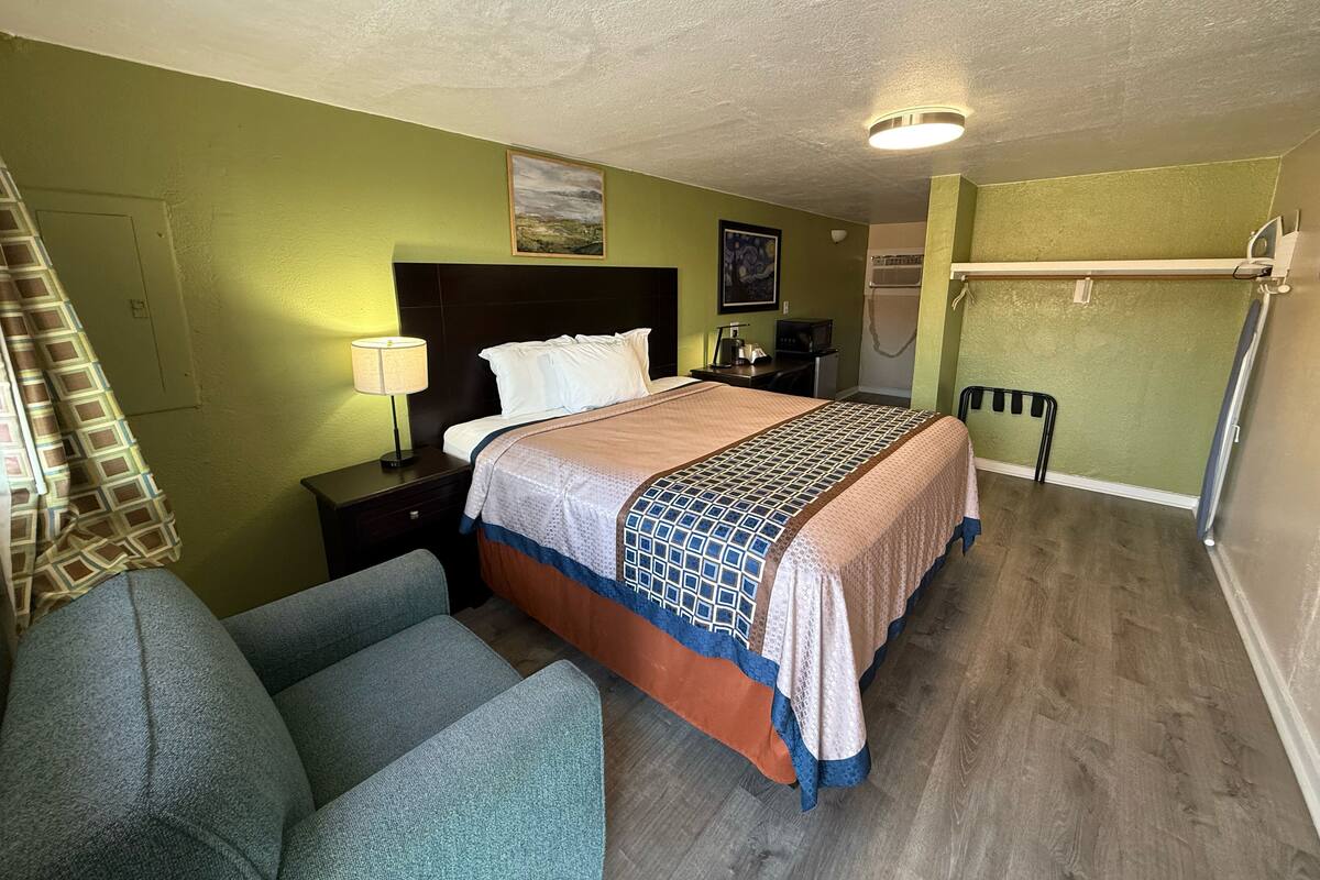 A hotel room with a bed, a desk, a chair, a lamp, a painting, and a shower area.