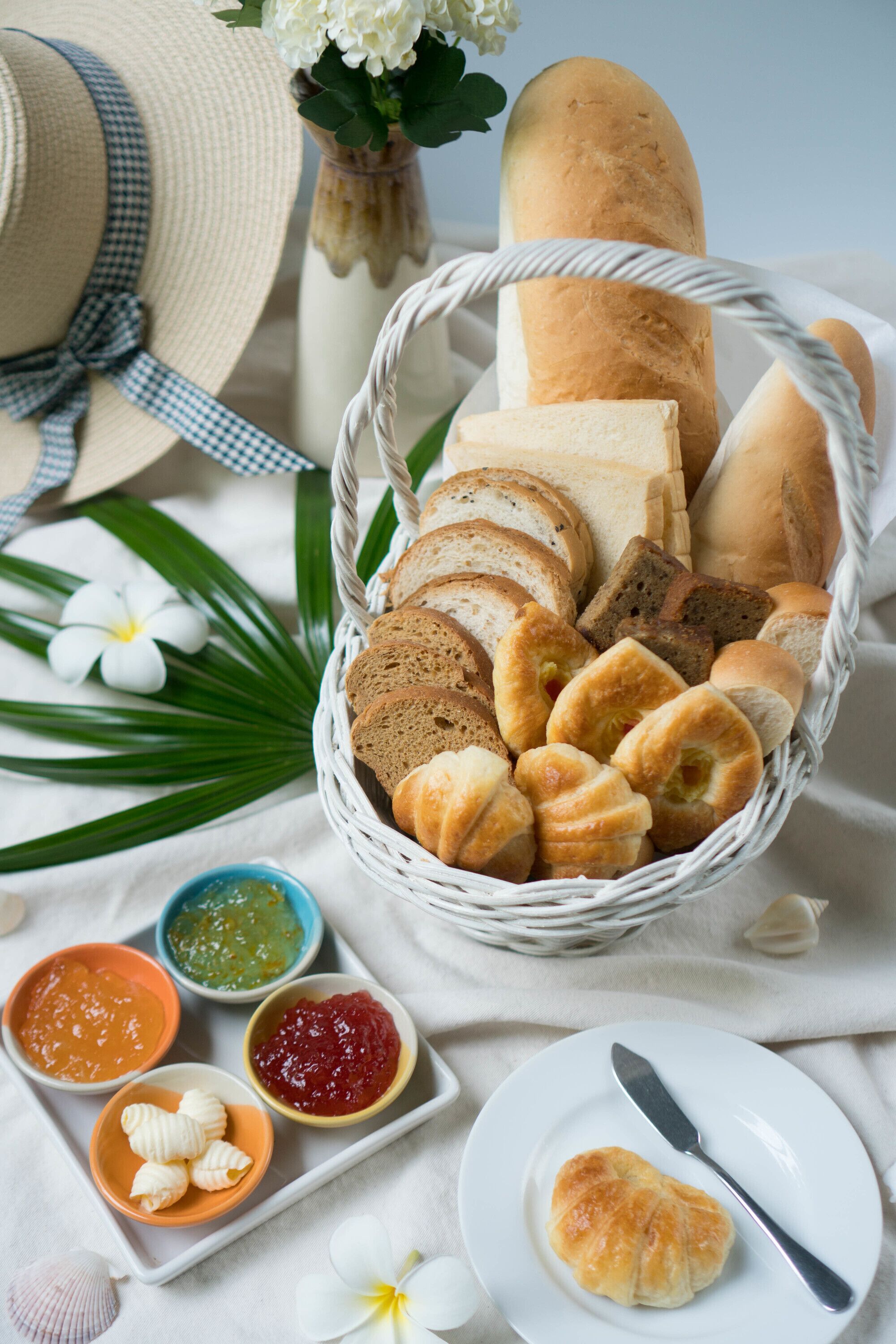 A basket of various breads and pastries, accompanied by small bowls of spreads and a white plate with a butter knife.