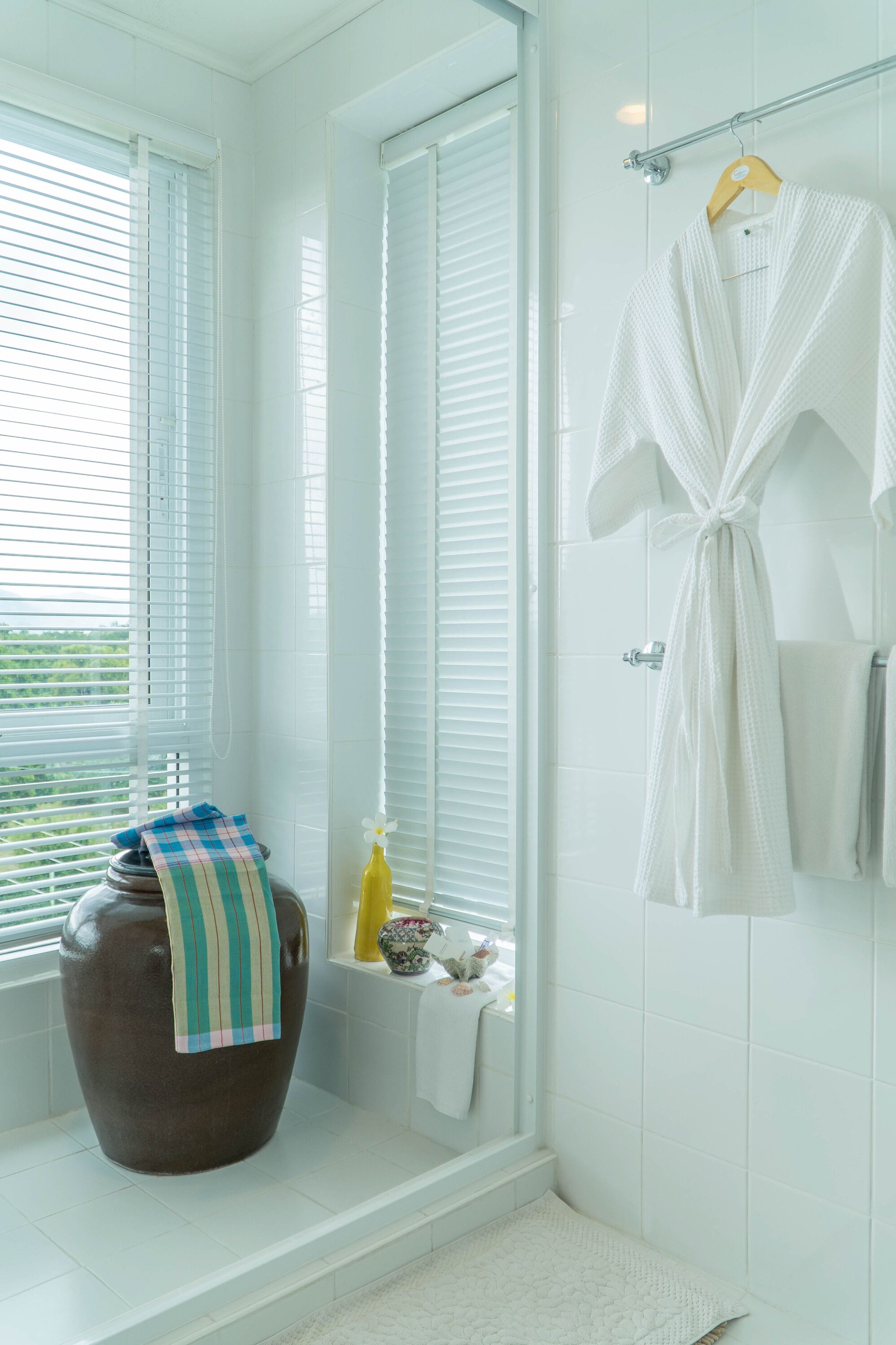 A bathroom with white tiles, a large window with blinds, a towel rack with a robe, a decorative vase, and a small shelf with toiletries.