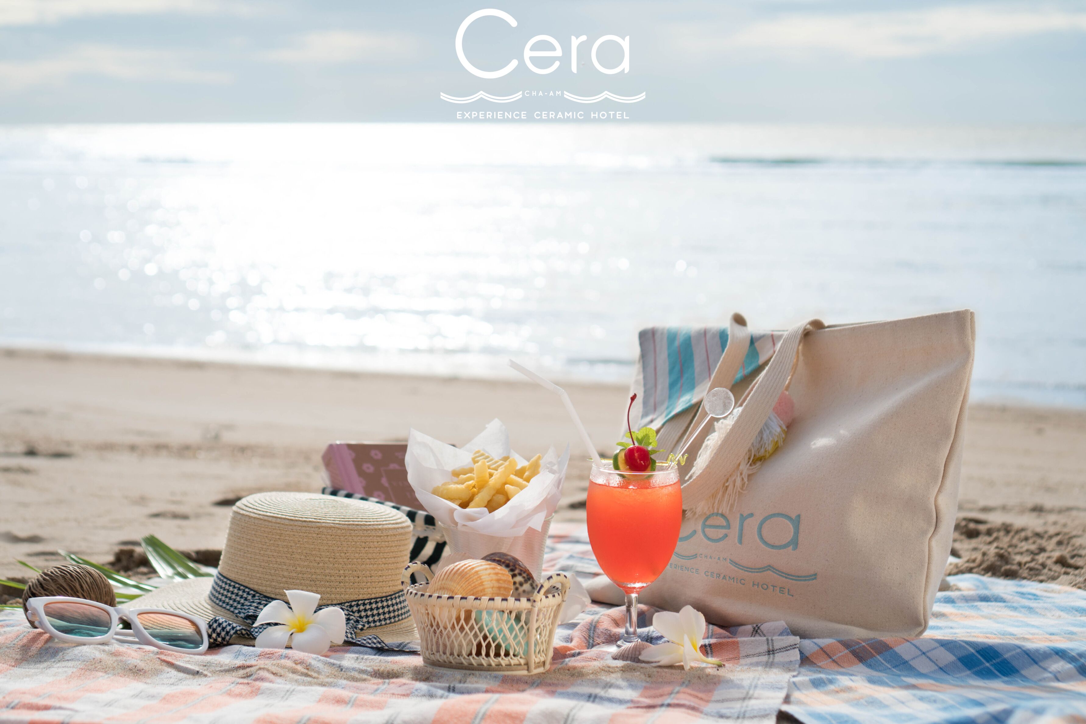 Escena de playa con un sombrero de paja, gafas de sol, una cesta de papas fritas, una bebida con cereza y un bolso de playa sobre una manta a cuadros.