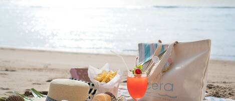 Escena de playa con un sombrero de paja, gafas de sol, una cesta de papas fritas, una bebida con cereza y un bolso de playa sobre una manta a cuadros.