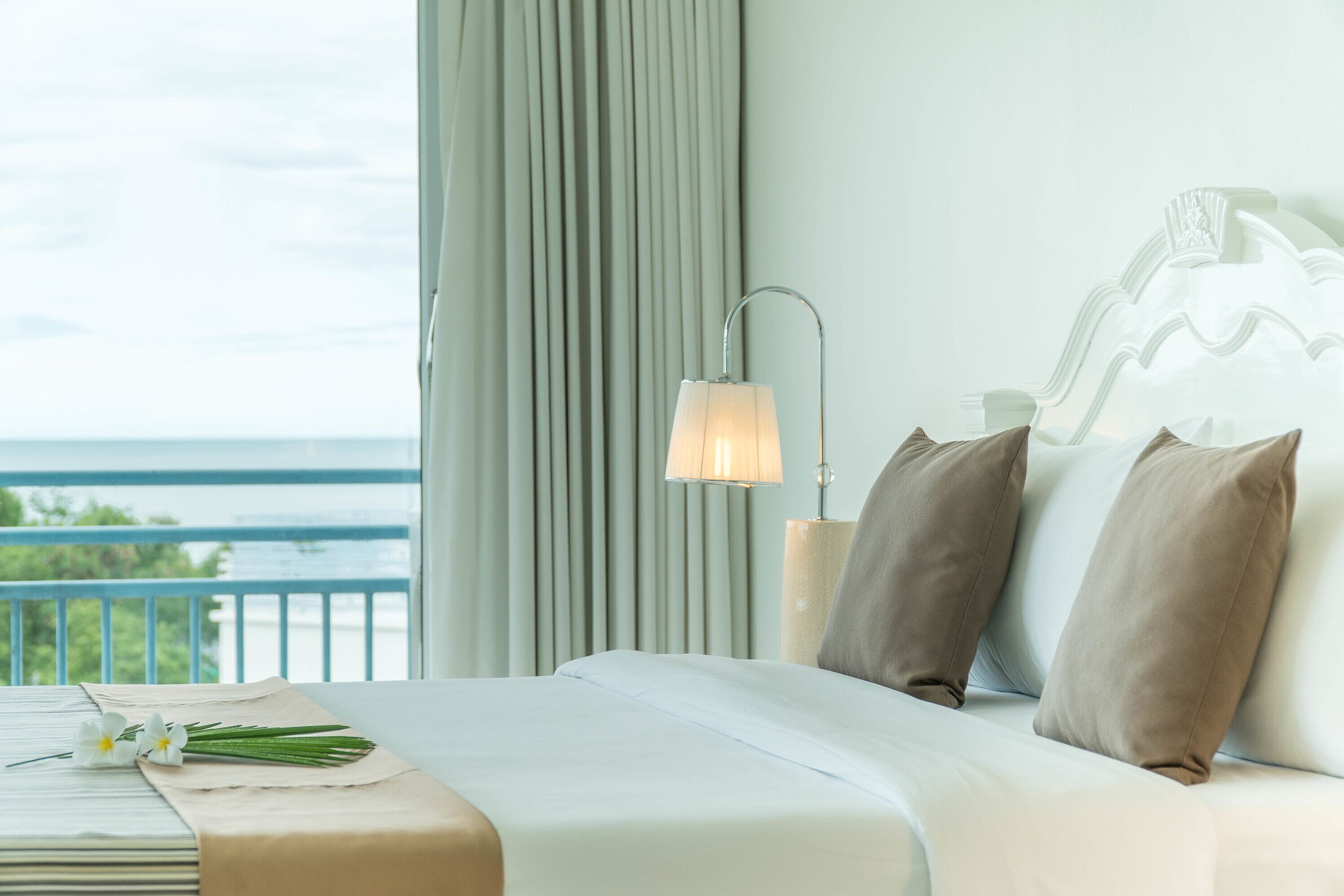 A neatly made bed with pillows, a bedside lamp, and a view of the ocean through curtains.