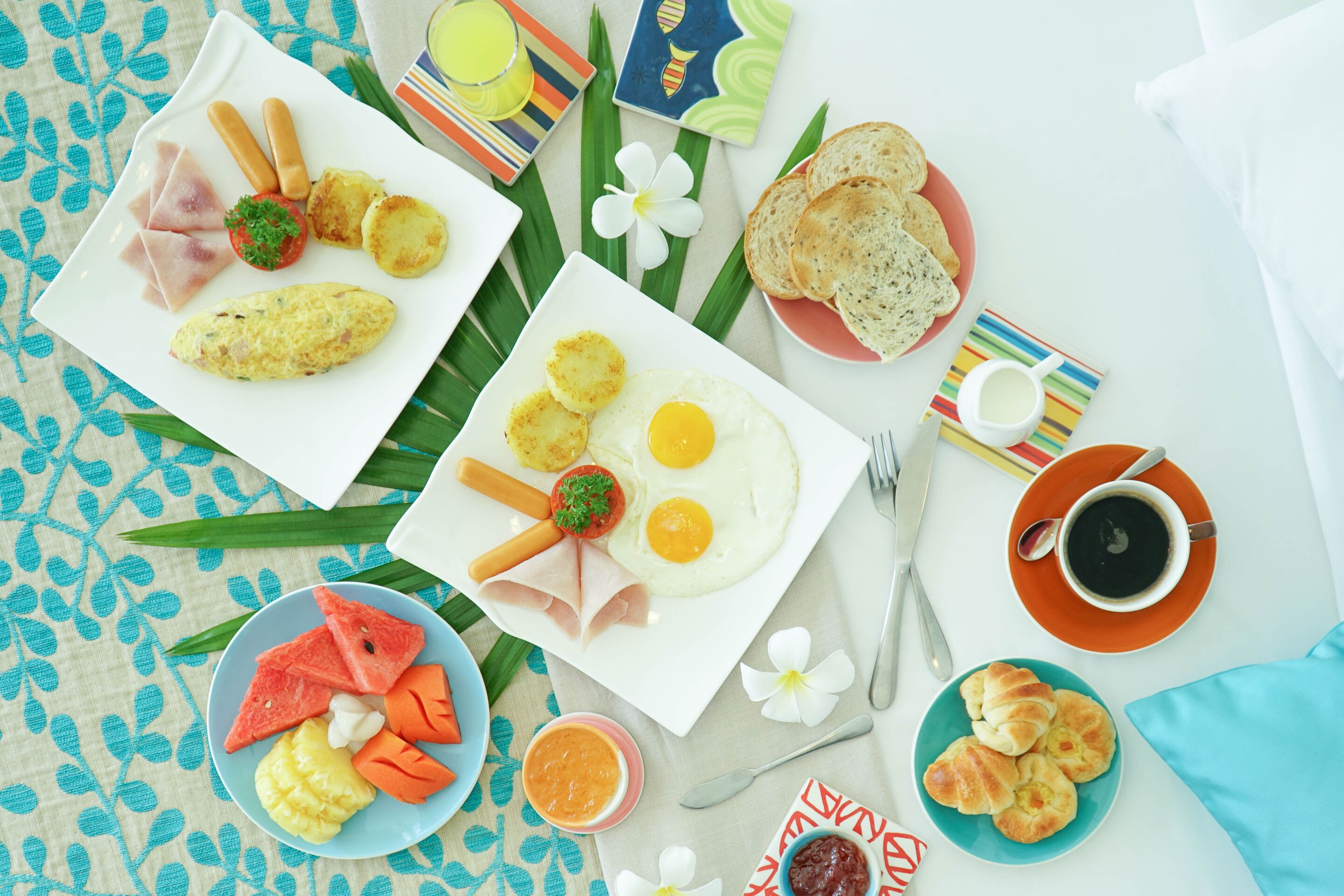 Un assortiment de plats pour le petit-déjeuner, comprenant des œufs, des toasts et des fruits, est disposé sur une table ornée d’un motif de feuilles tropicales.