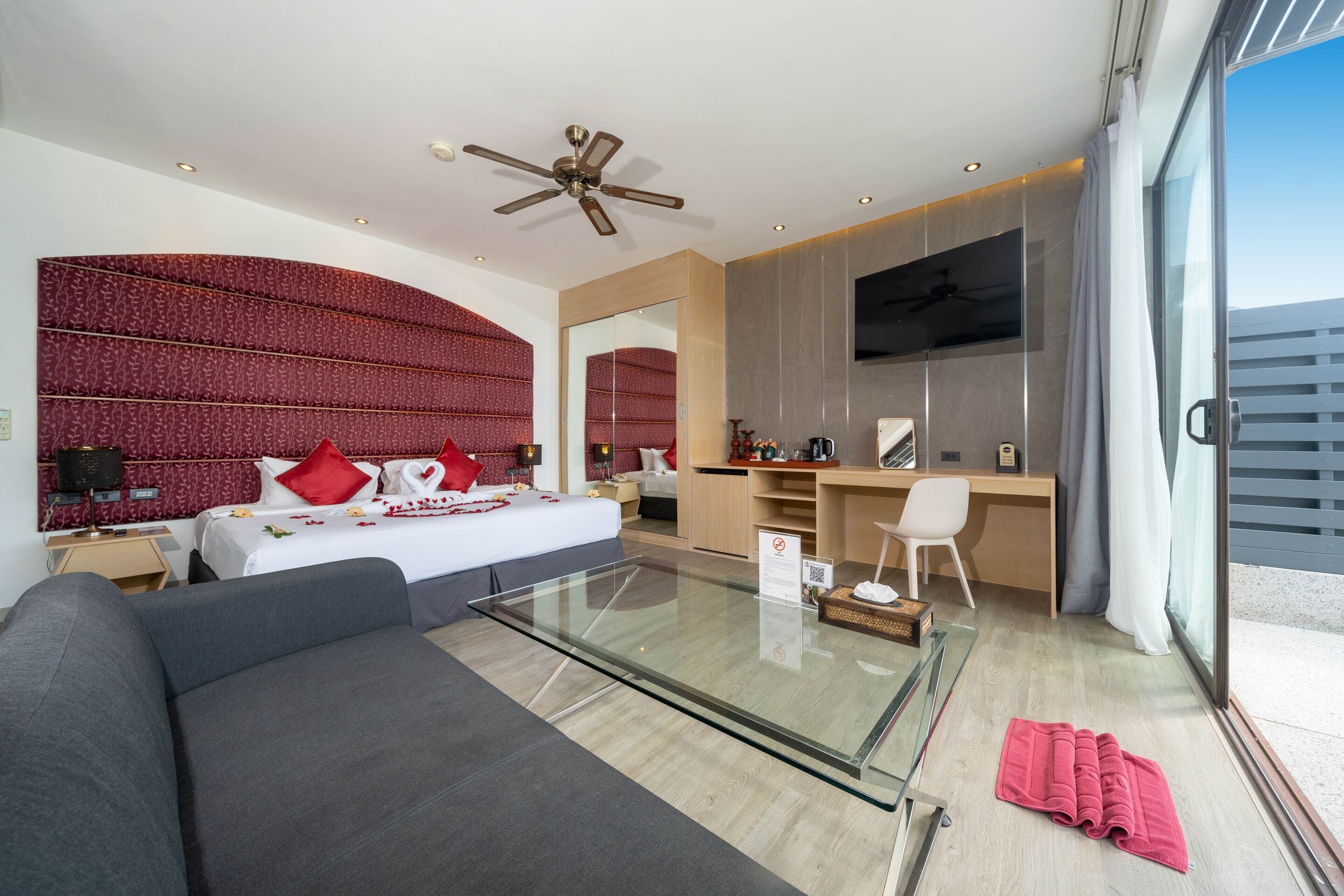 A modern hotel room with a glass coffee table, a bed with red pillows, a desk with a chair, and a large mirror.