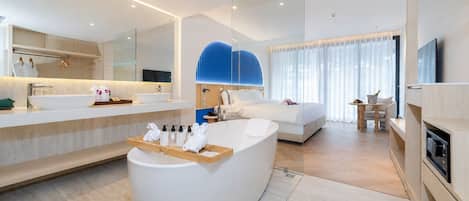 A modern hotel room with a large bathtub, a separate shower area, a bed, a desk, and a television.