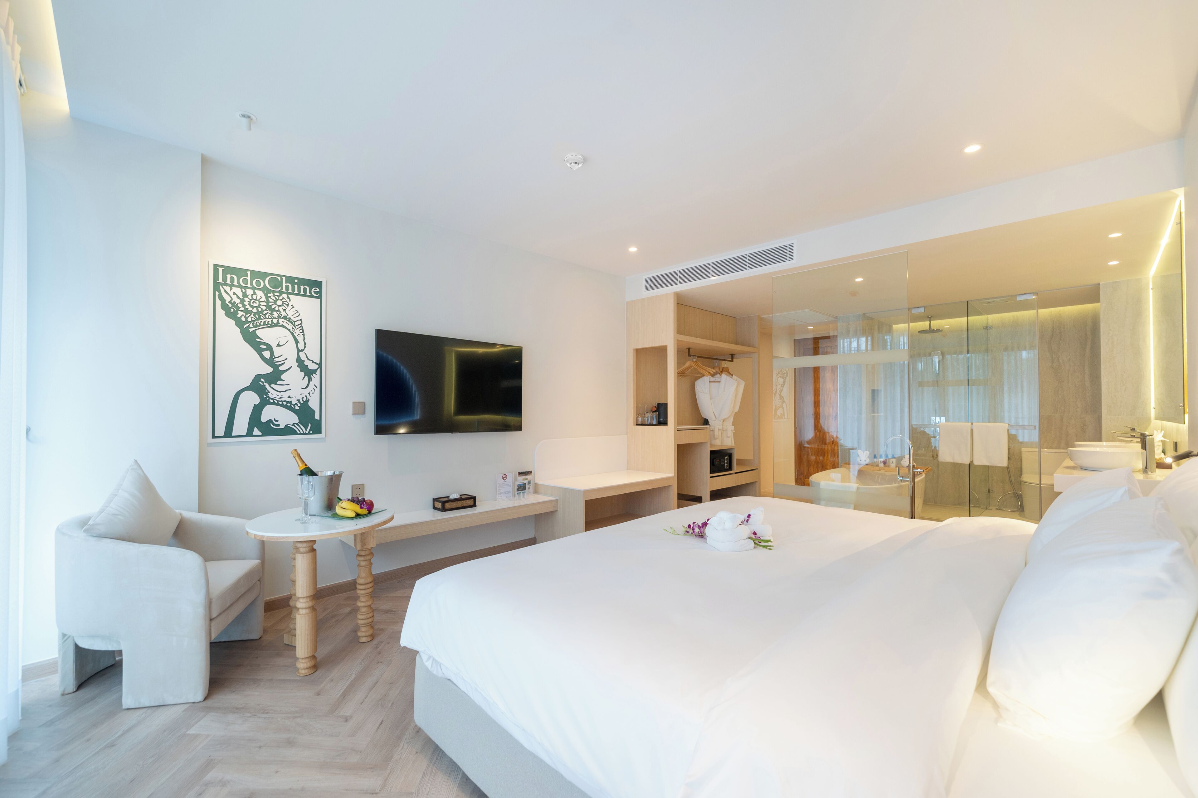 A modern hotel room with a large bed, a seating area with a table, a TV, a mirror, and a wardrobe.