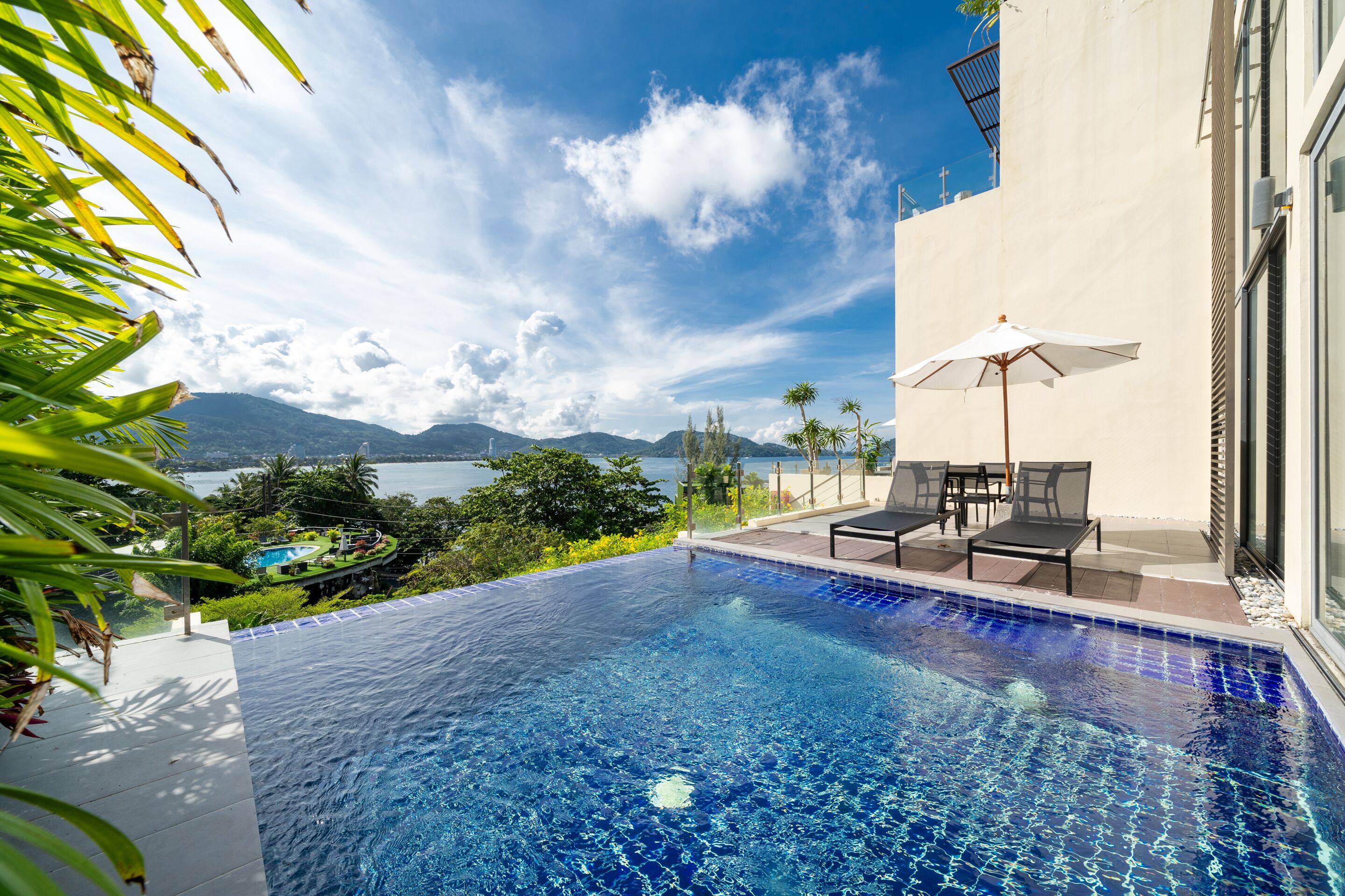 A clear blue infinity pool with a view of mountains and a body of water. There are lounge chairs and an umbrella on the patio.