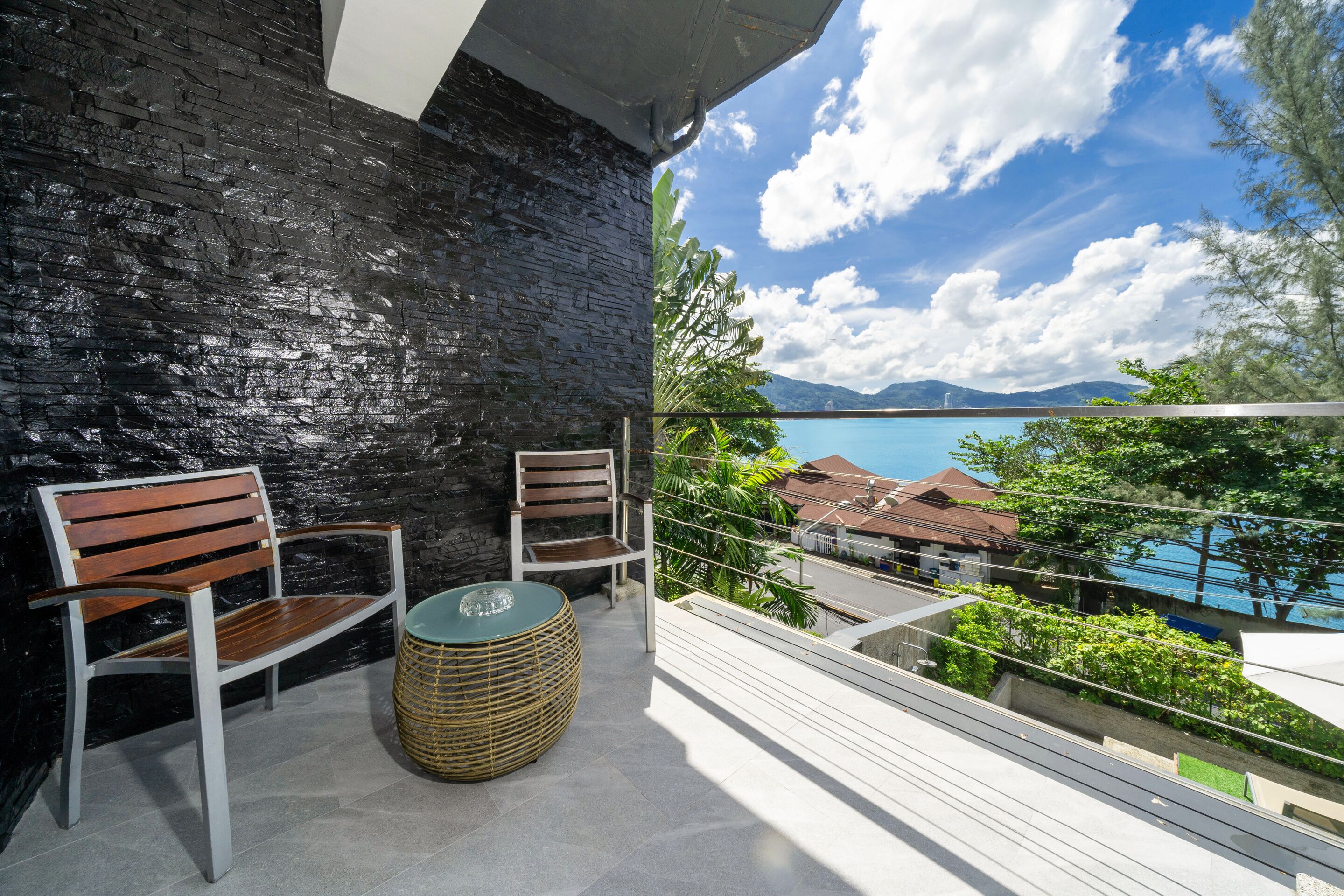 A balcony with two wooden chairs and a woven stool, overlooking a body of water and a mountainous landscape.
