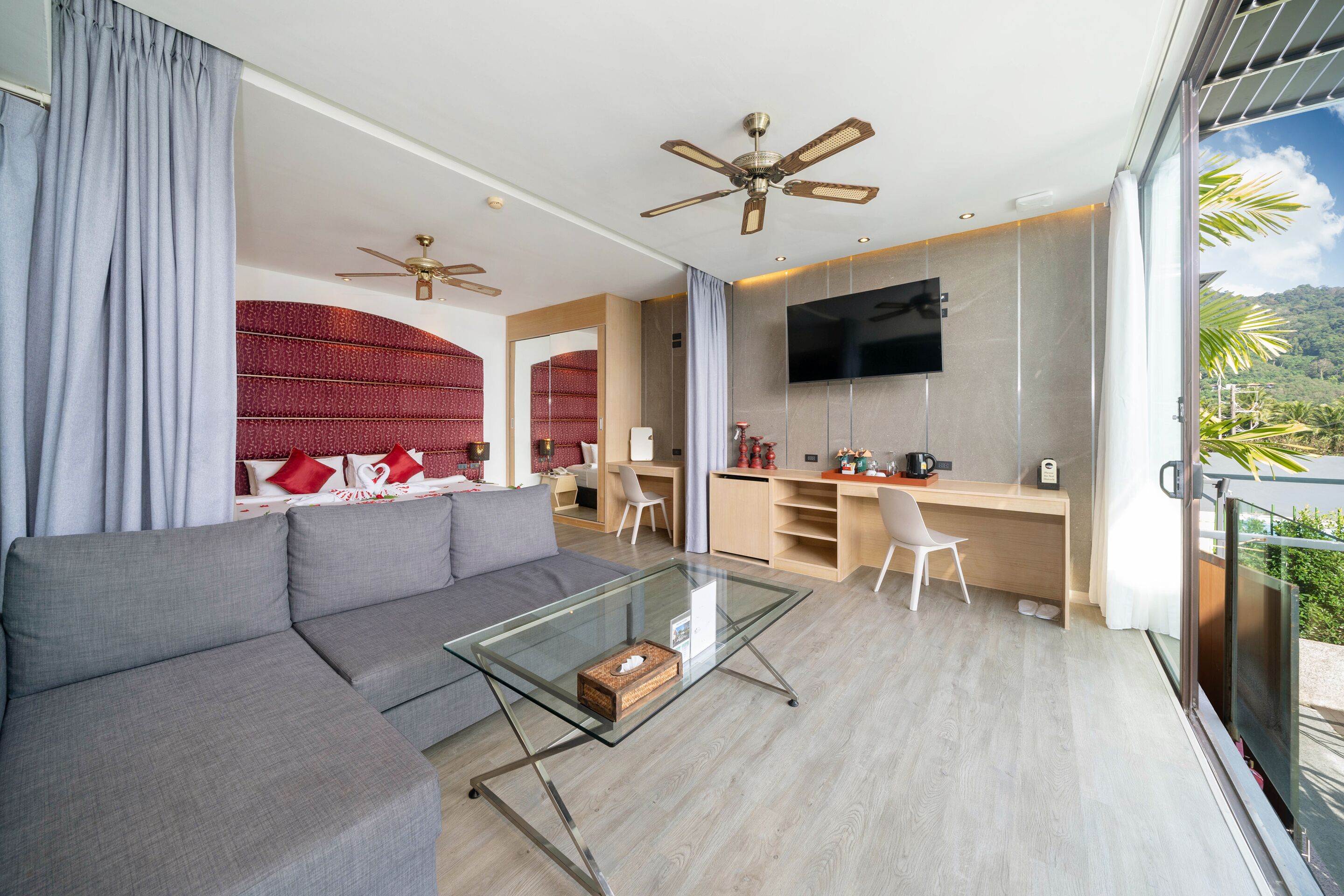 A modern living room with a gray sofa, a glass coffee table, and a TV mounted on the wall. There is a kitchen area with a wooden countertop and a chair.