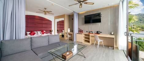 A modern living room with a gray sofa, a glass coffee table, and a TV mounted on the wall. There is a kitchen area with a wooden countertop and a chair.