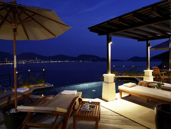 An outdoor pool area with lounge chairs and a table, overlooking a body of water at night.