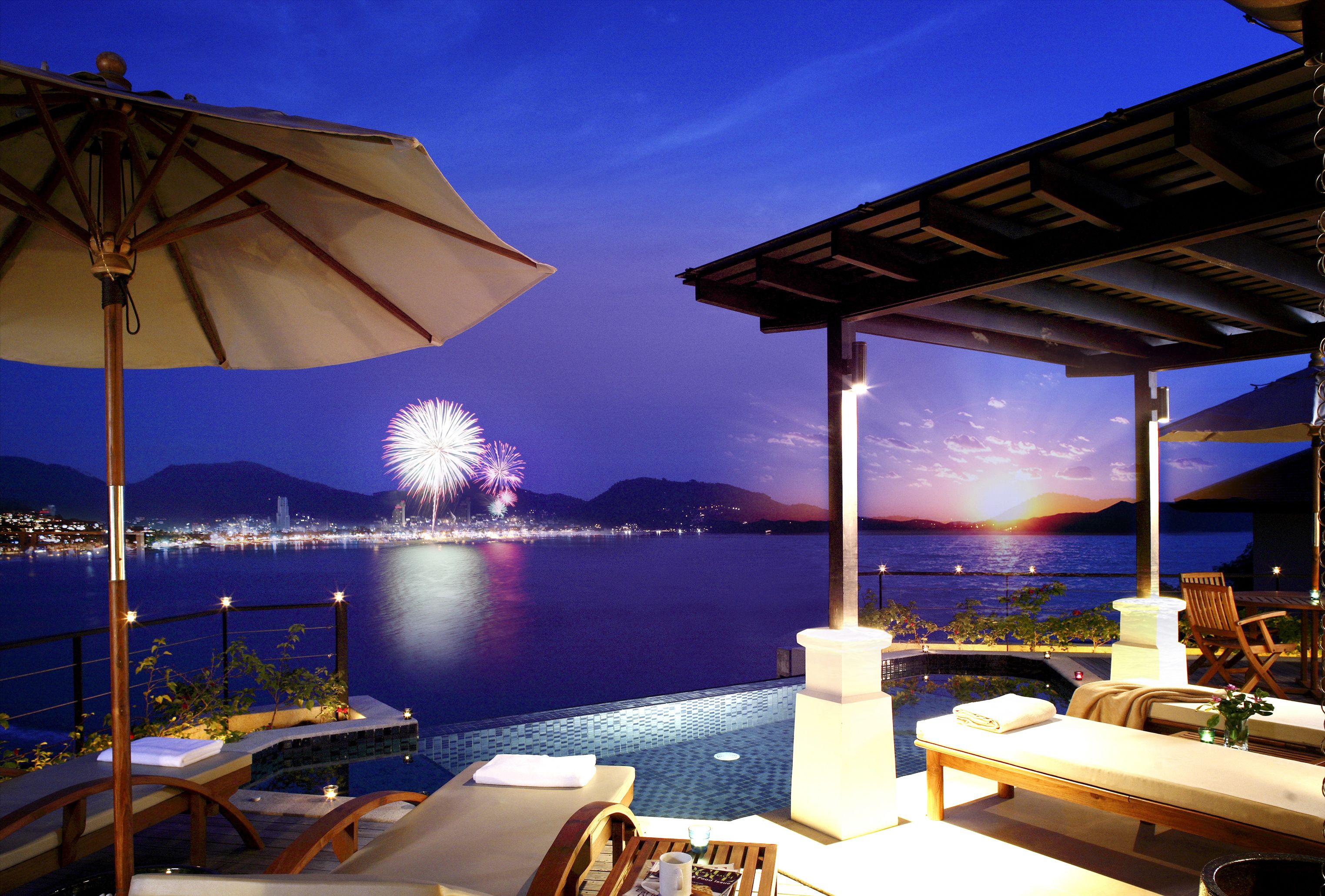 A poolside area with lounge chairs, an umbrella, and a view of the water with fireworks in the background.