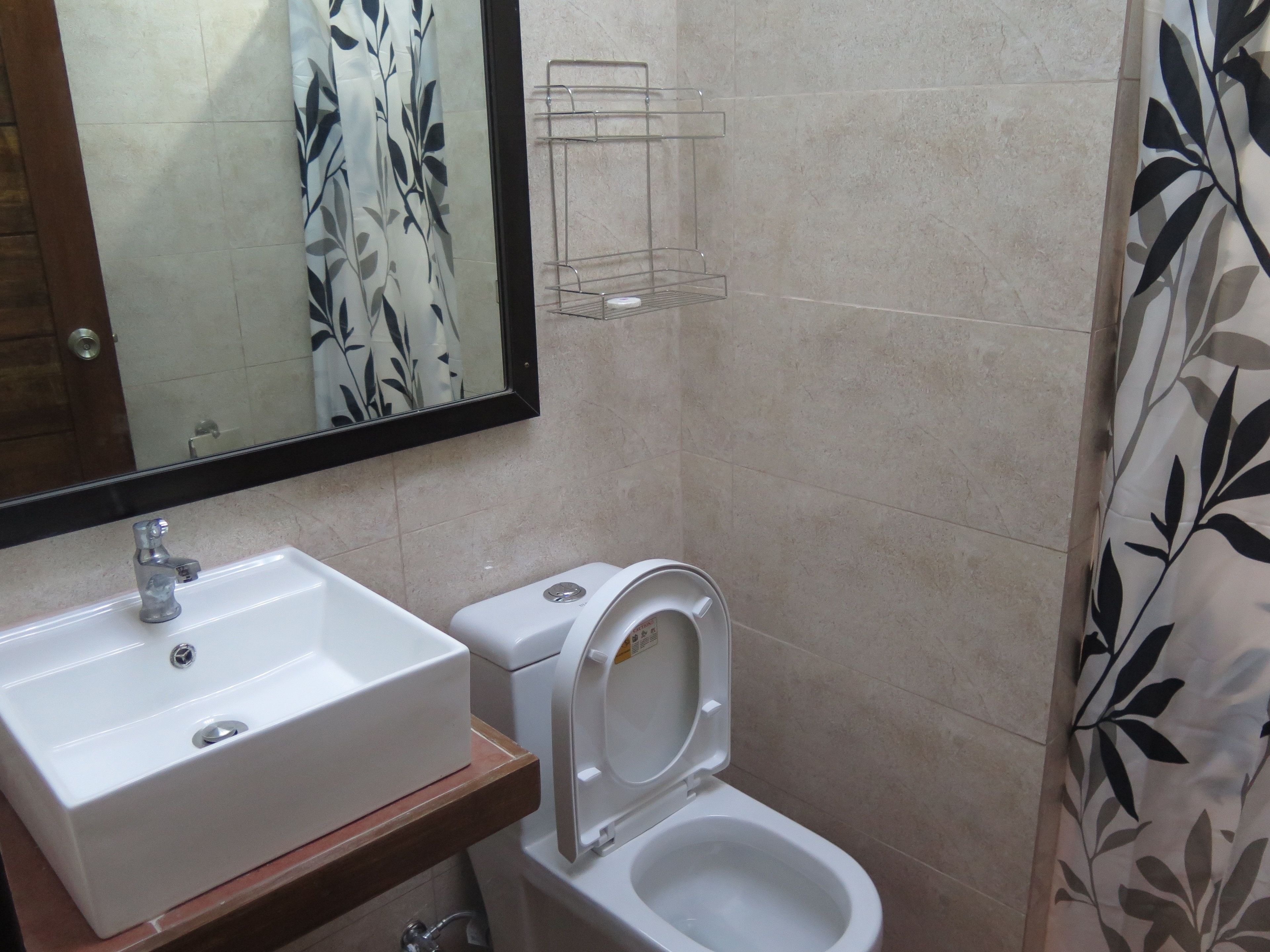 Une salle de bain avec un lavabo blanc, des toilettes et un rideau de douche orné d’un motif de feuilles.