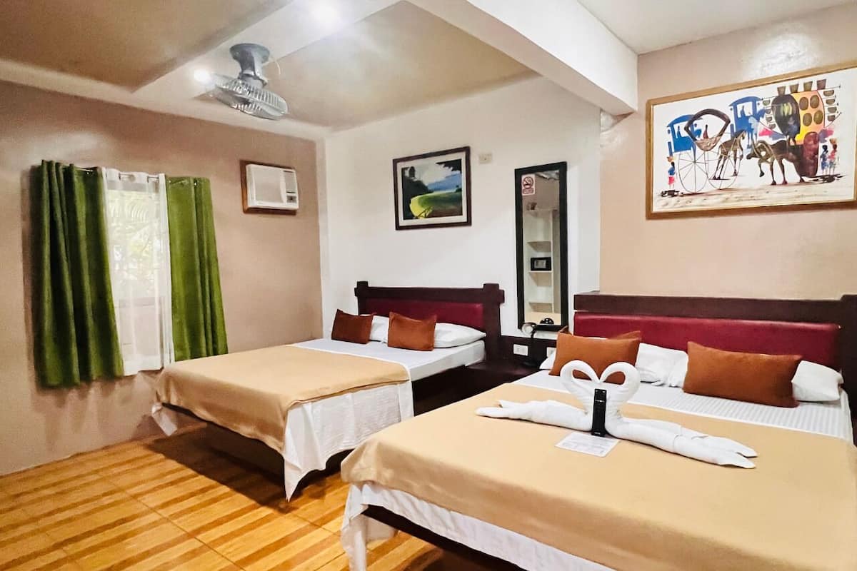 Une chambre d’hôtel avec deux lits, un plancher en bois, un ventilateur au plafond, un tableau au mur et une fenêtre avec des rideaux verts.
